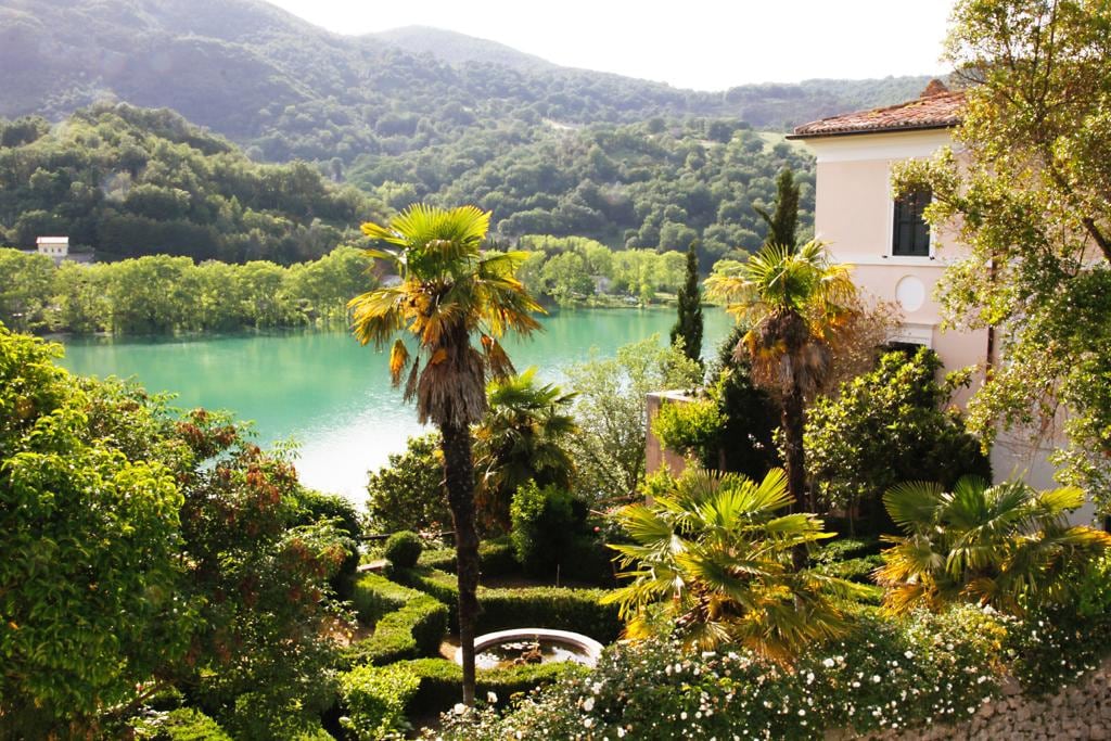 The image captures a lush garden filled with palm trees and vibrant greenery, overlooking the serene turquoise waters of the lake. Rolling hills and a forested backdrop provide a tranquil setting, while a fountain is partially visible within the garden.