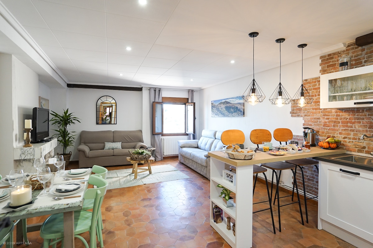 The spacious living area features a modern kitchen bar with wooden stools and a dining table set for four. A comfortable sofa and a second seating area are visible, with natural light streaming through windows. Exposed brick accents provide character to the room.