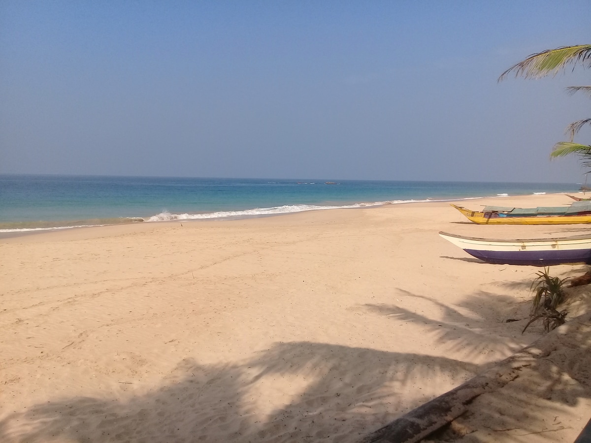 A spacious stretch of unspoiled beach is visible, featuring soft, golden sand meeting calm, turquoise waters. A few traditional fishing boats are positioned along the shore, while gentle waves lapping at the sand create a serene backdrop under a clear sky.