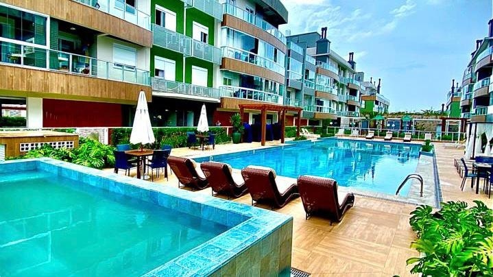 A spacious outdoor swimming area features a large pool surrounded by lounge chairs. A heated jacuzzi is visible nearby, and an area with umbrellas and tables is present. Modern buildings with balconies enclose the space, creating a welcoming atmosphere for relaxation and leisure.