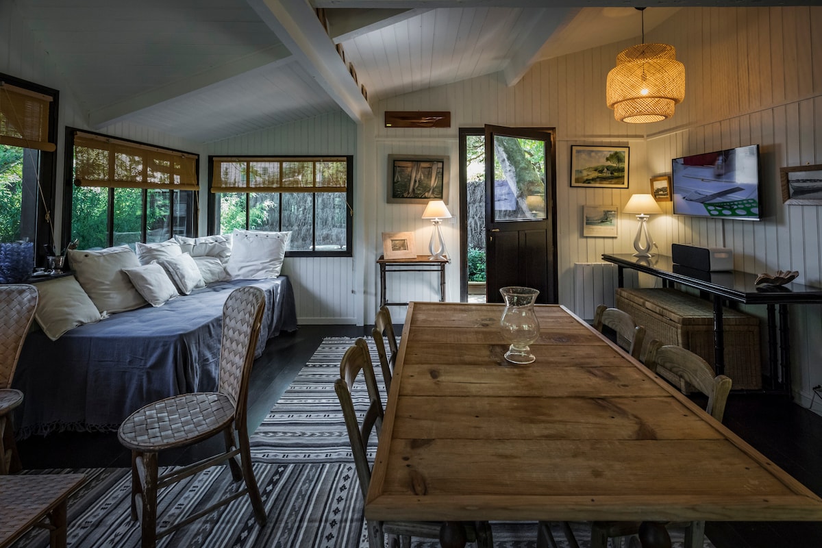 A cozy interior space features a wooden dining table with several chairs, complemented by a relaxed seating area adorned with plush cushions. Natural light illuminates the room, highlighting the wooden accents and soft textiles against a backdrop of greenery visible through large windows.