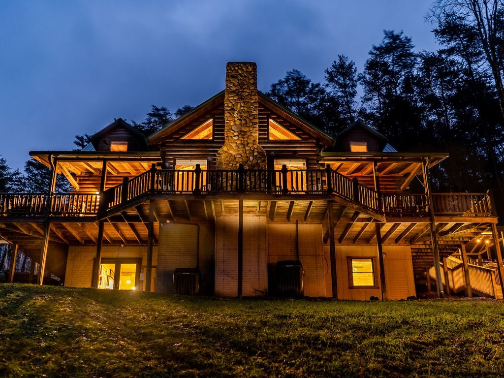 The exterior of The Lodge is illuminated at dusk, showcasing a spacious structure with a large stone chimney at the center. The wraparound deck provides ample seating and invites guests to enjoy the surrounding landscape. Warm light spills from multiple windows, enhancing the inviting appearance.