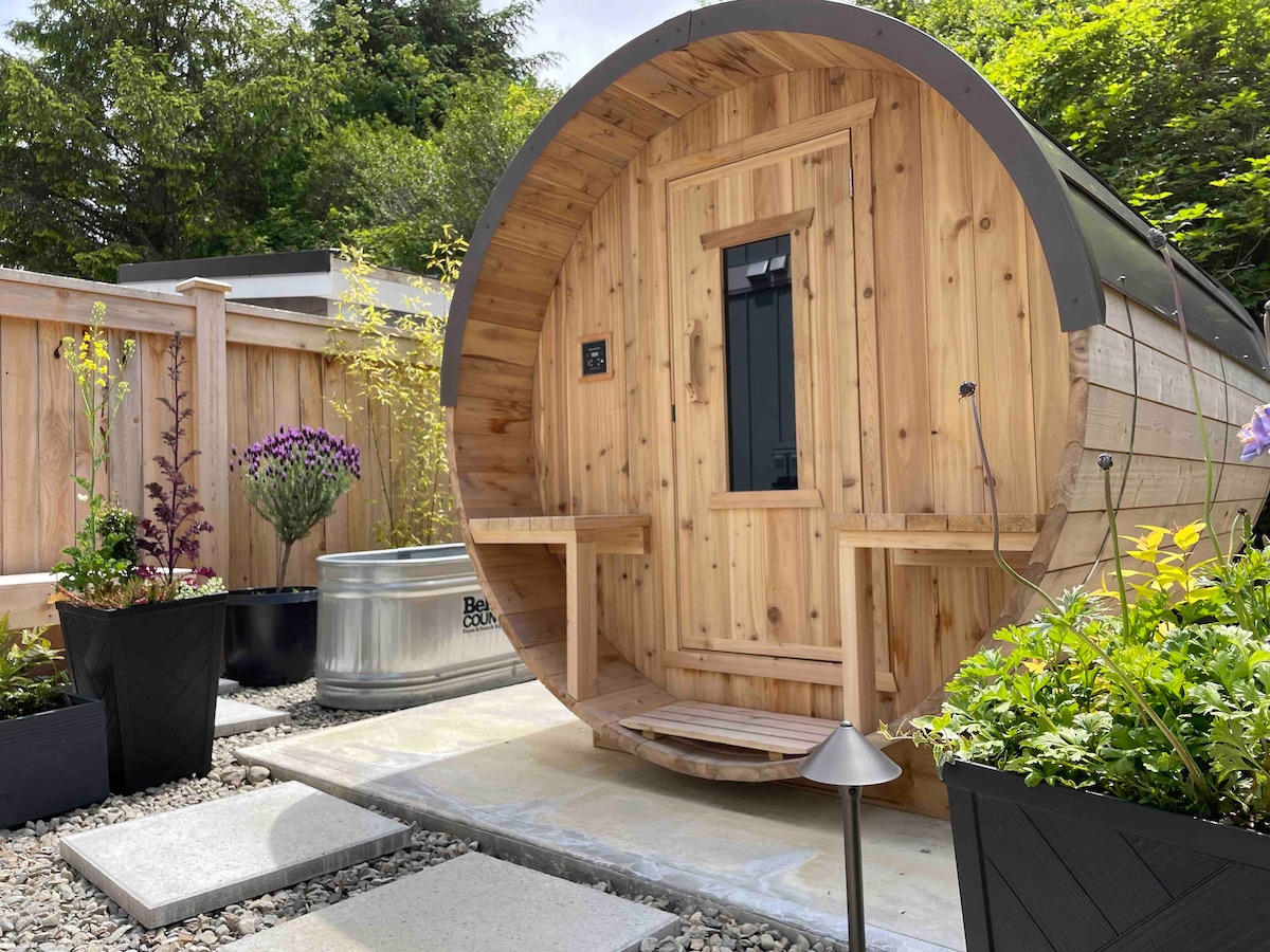 The outdoor space features a circular wooden sauna with a door and two benches designed for relaxation. Surrounding the sauna are neatly arranged flower pots and a stone pathway leading to it, contributing to a serene garden environment.