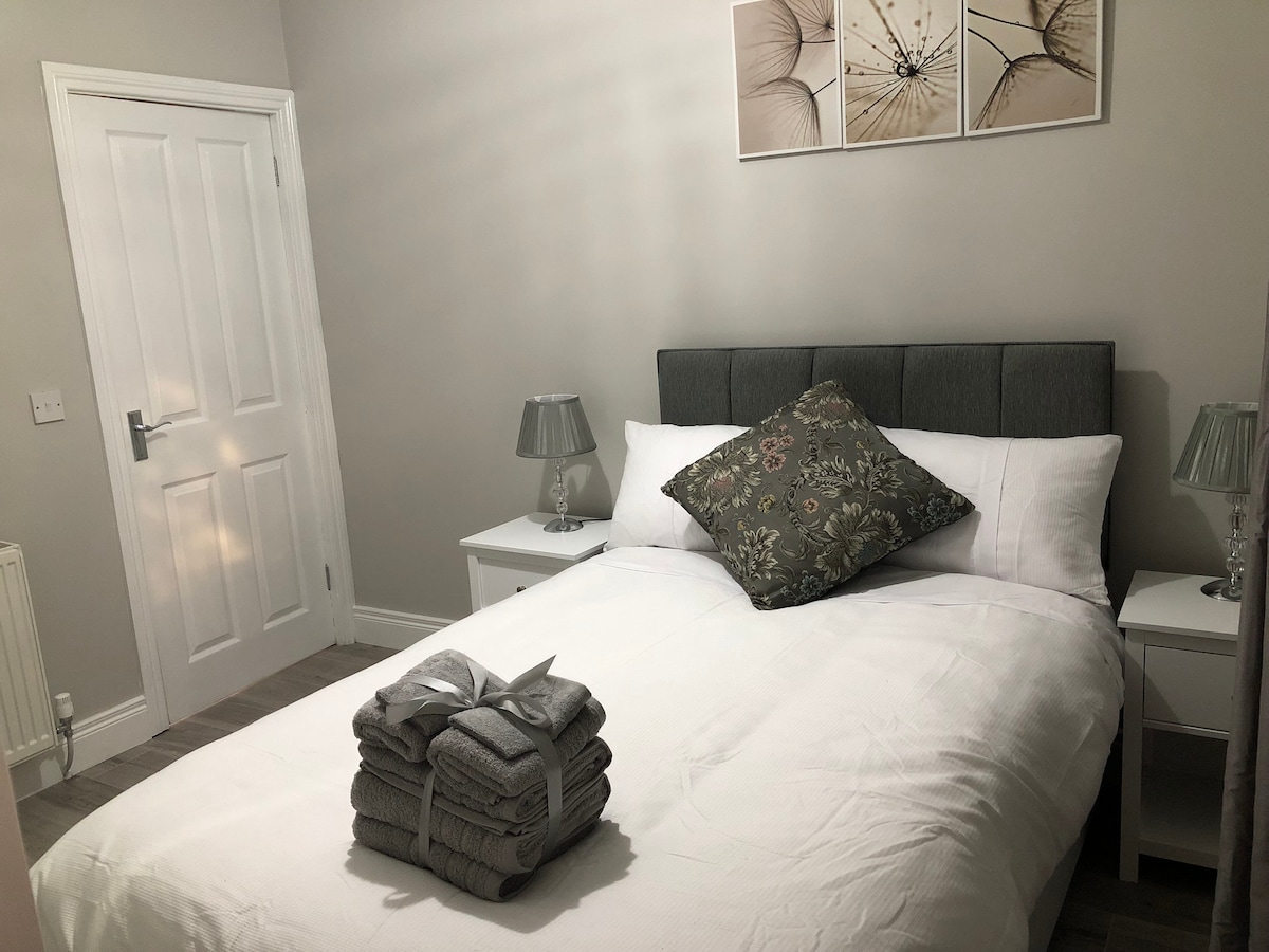 A small double bed is neatly made with white linens and a decorative pillow. On either side of the bed are light-colored nightstands with table lamps. A stack of grey towels is displayed at the foot of the bed, and floral artwork adorns the wall above.