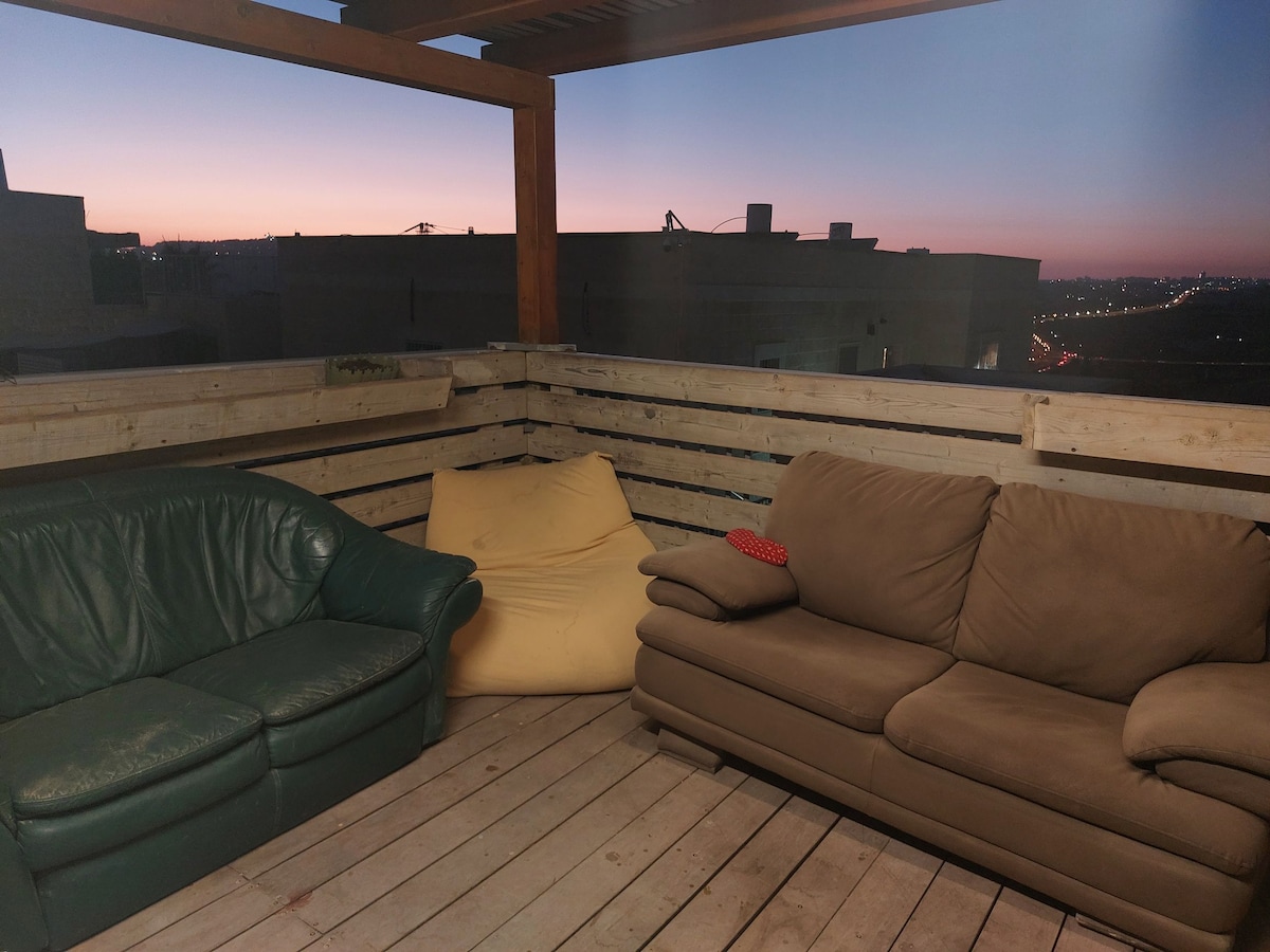 A wooden deck is surrounded by a fence, featuring two couches in contrasting colors, one green and one beige, alongside a yellow bean bag. The evening sky is visible in the background, transitioning to twilight, with urban scenery beyond the balcony.