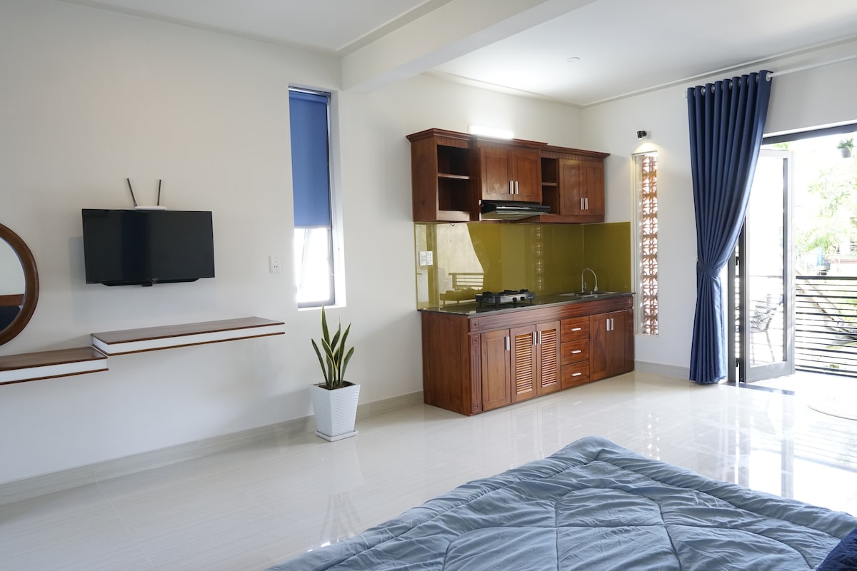 A spacious apartment interior is showcased, featuring a modern kitchenette with wooden cabinetry and a small dining area. The room is well lit by natural daylight from large windows, with a cozy sleeping area visible. Decorative plants add a touch of greenery.