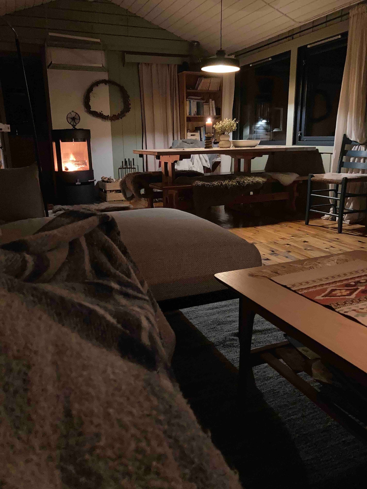 A cozy interior is illuminated by warm light from a wood stove. A dining table is set in the background, accompanied by chairs and a bench seating area. Soft textures from blankets and rugs create a comfortable atmosphere in the spacious room.