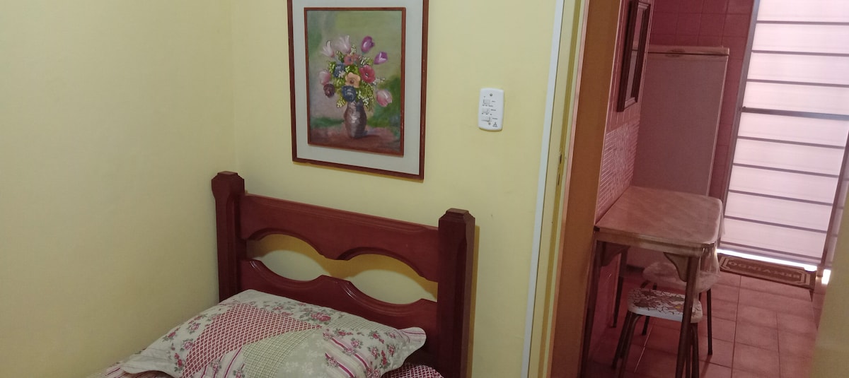 A small bedroom is depicted, featuring a wooden bed with a quilted coverlet. A floral painting hangs on the yellow wall, adding a touch of color. In the background, a simple dining table is visible, situated near the entrance to a small kitchen.