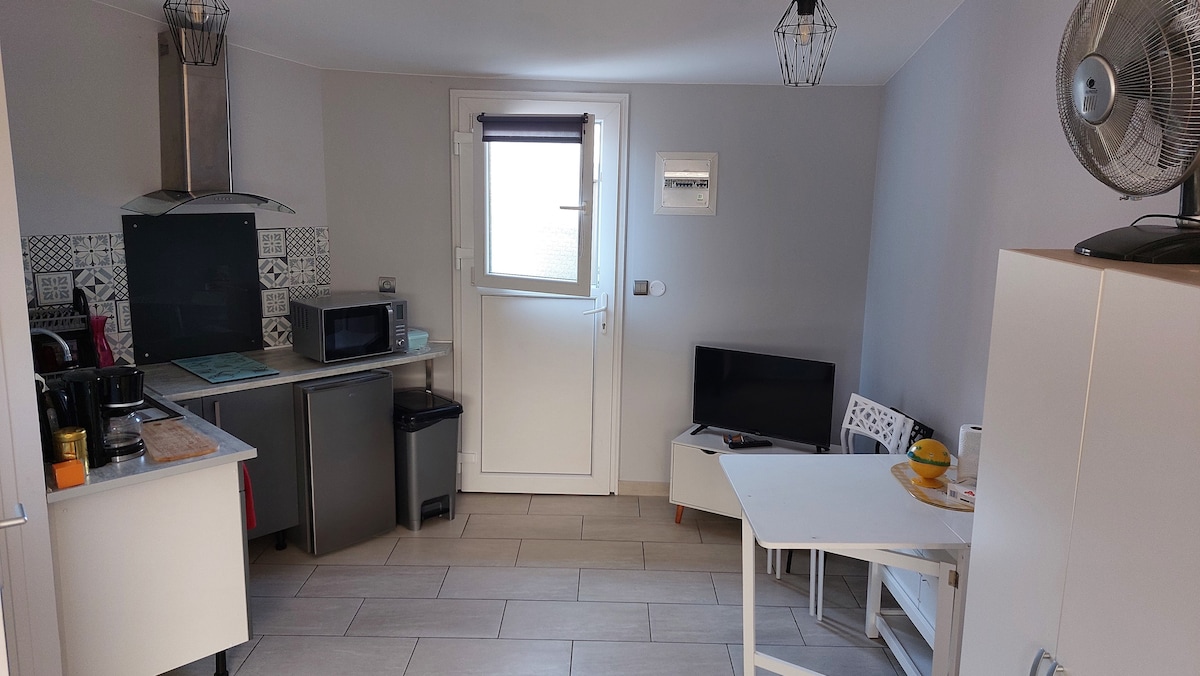 The kitchen area features modern appliances including a microwave and coffee maker, with ample countertop space. A small white table and chairs are positioned beside a television. Natural light enters through a window, and the door leads to the outside.
