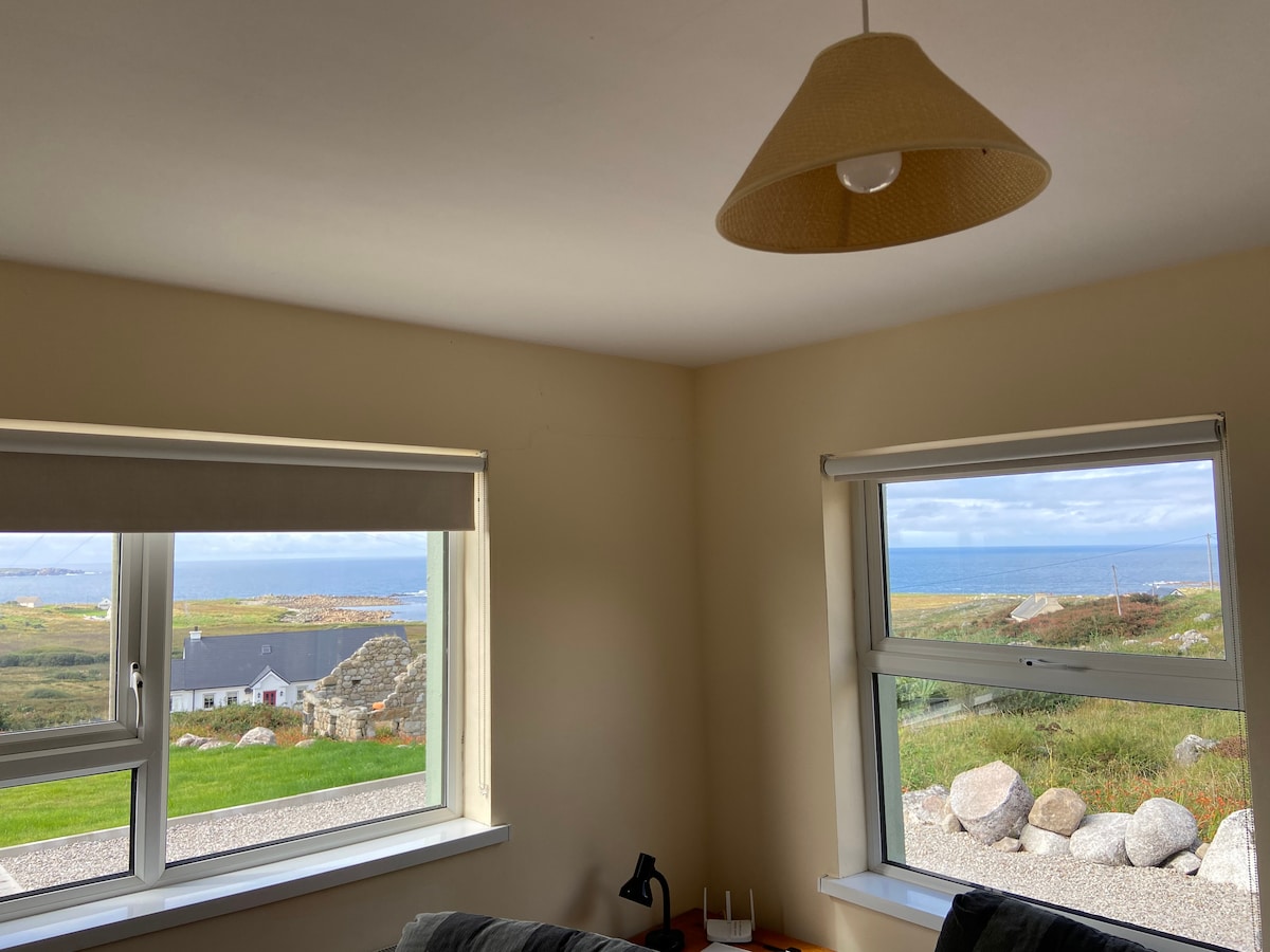Two large windows offer scenic views of the Atlantic Ocean and surrounding landscape. Natural light fills the room, highlighting neutral-colored walls. A minimalist lamp and a small table are visible, contributing to a peaceful and functional living space.