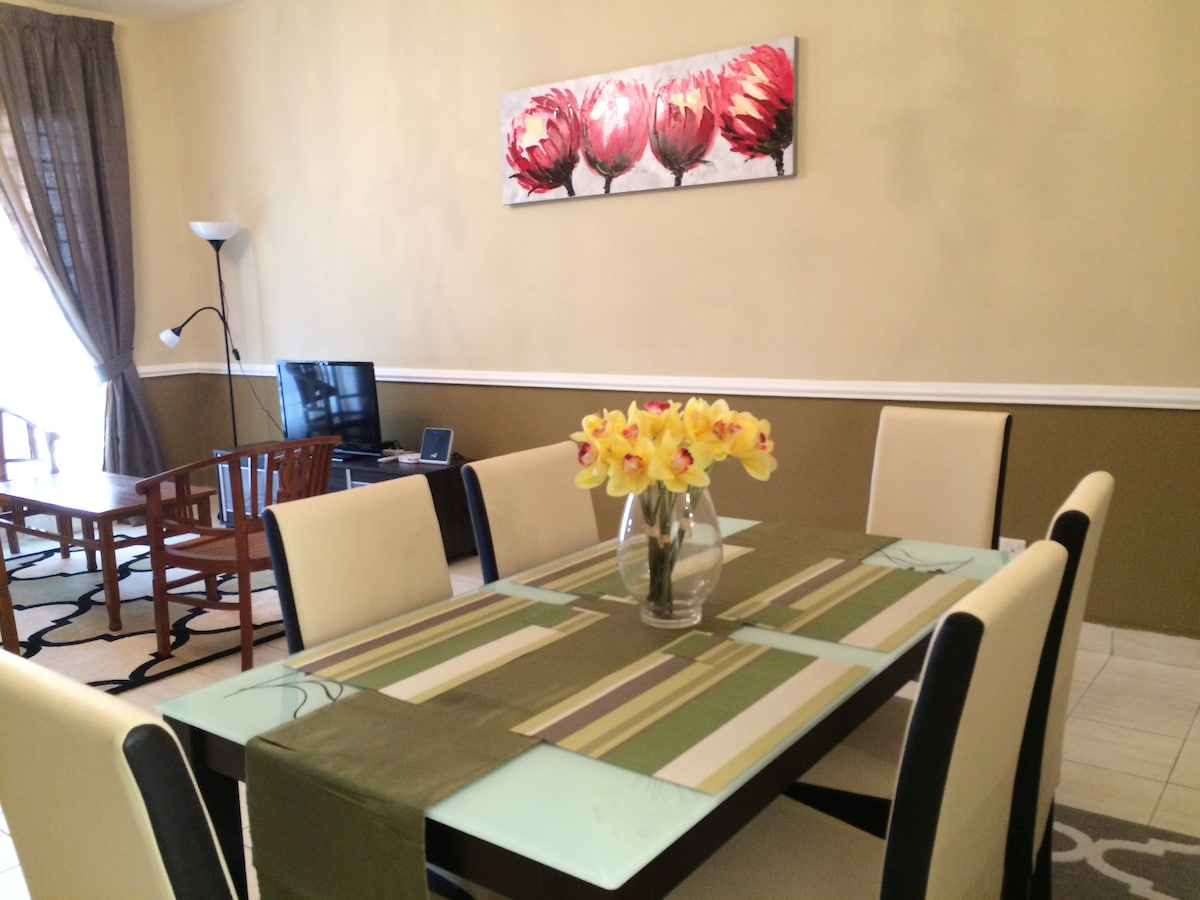 A dining area features a rectangular table surrounded by six light-colored chairs. A vase of yellow flowers sits at the center. A large artwork depicting red flowers is displayed on the wall above the table. Natural light enters through a nearby window, framed by dark curtains.