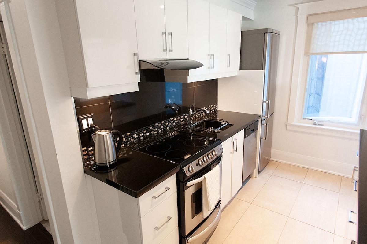 The modern kitchen features sleek white cabinetry paired with a dark countertop. Stainless steel appliances include a stove, refrigerator, and a kettle, providing a contemporary look. A window allows natural light, highlighting the clean tile flooring throughout the space.