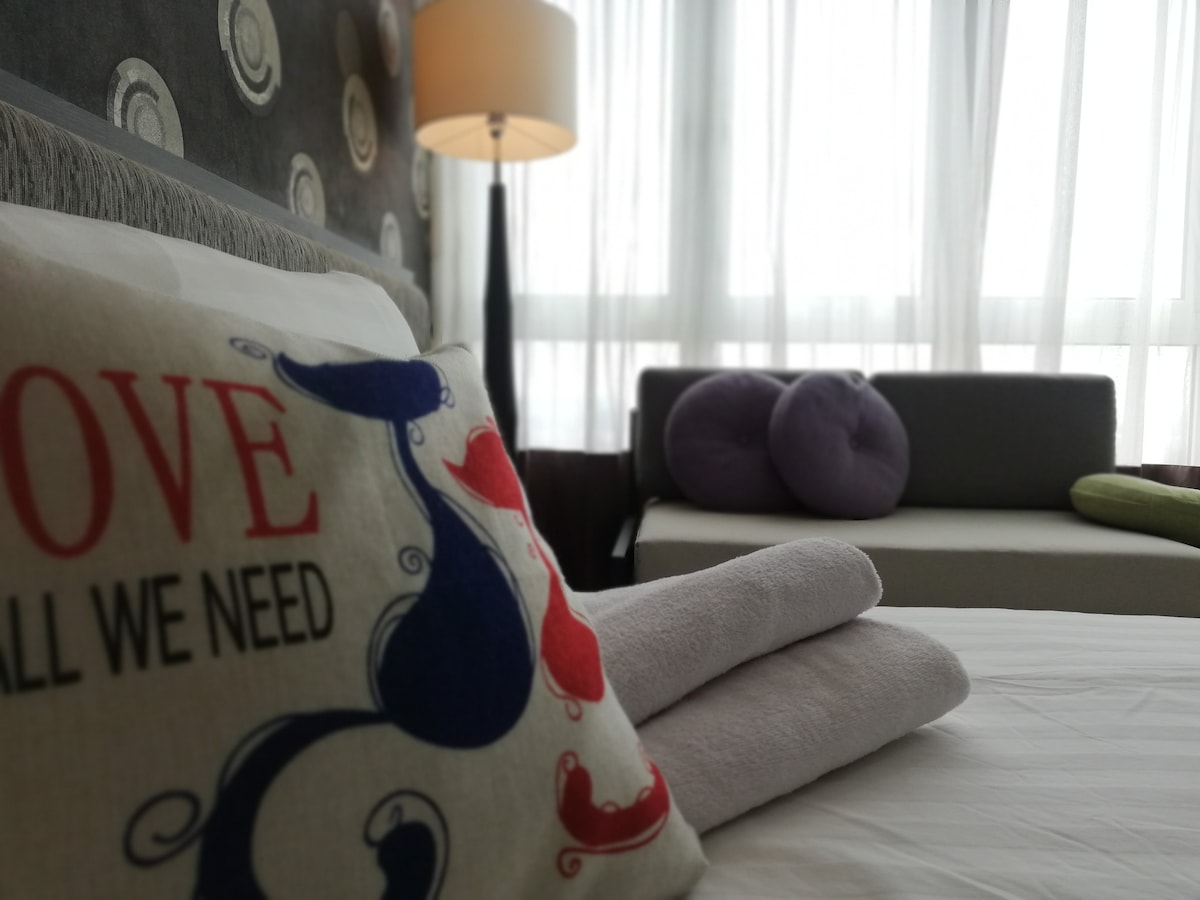 A cozy bedroom scene features a spacious bed adorned with a decorative pillow showcasing a simple message. Soft towels are neatly arranged nearby. In the background, a sofá is visible, alongside a lamp casting gentle light through sheer curtains that allow natural light to filter in.
