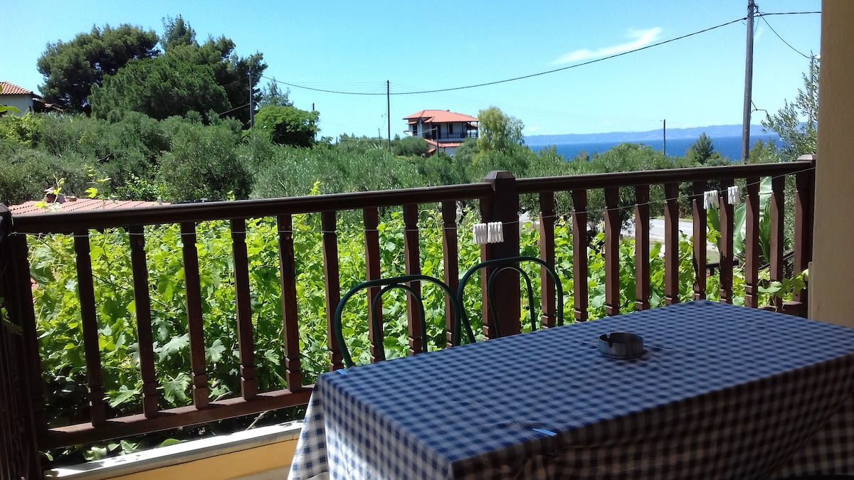 A spacious balcony showcases a checkered tablecloth-covered table, paired with two chairs. Lush greenery lines the view, with trees and distant buildings visible against a backdrop of blue sea and clear sky.