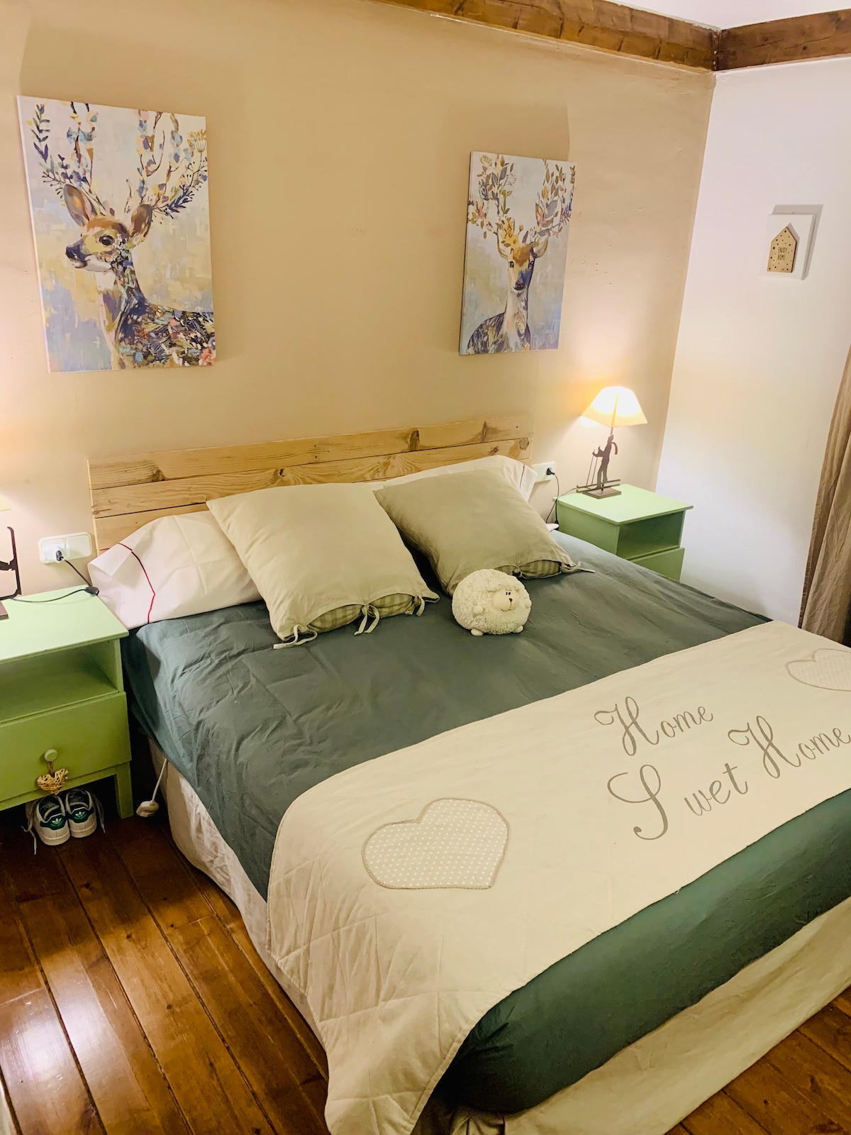 A cozy bedroom features a spacious bed with soft linens and two bedside tables. Artwork depicting deer adorns the wall above, complemented by gentle lighting from a lamp on one side. A decorative blanket with 'Home Sweet Home' is displayed across the bed, enhancing the inviting atmosphere.