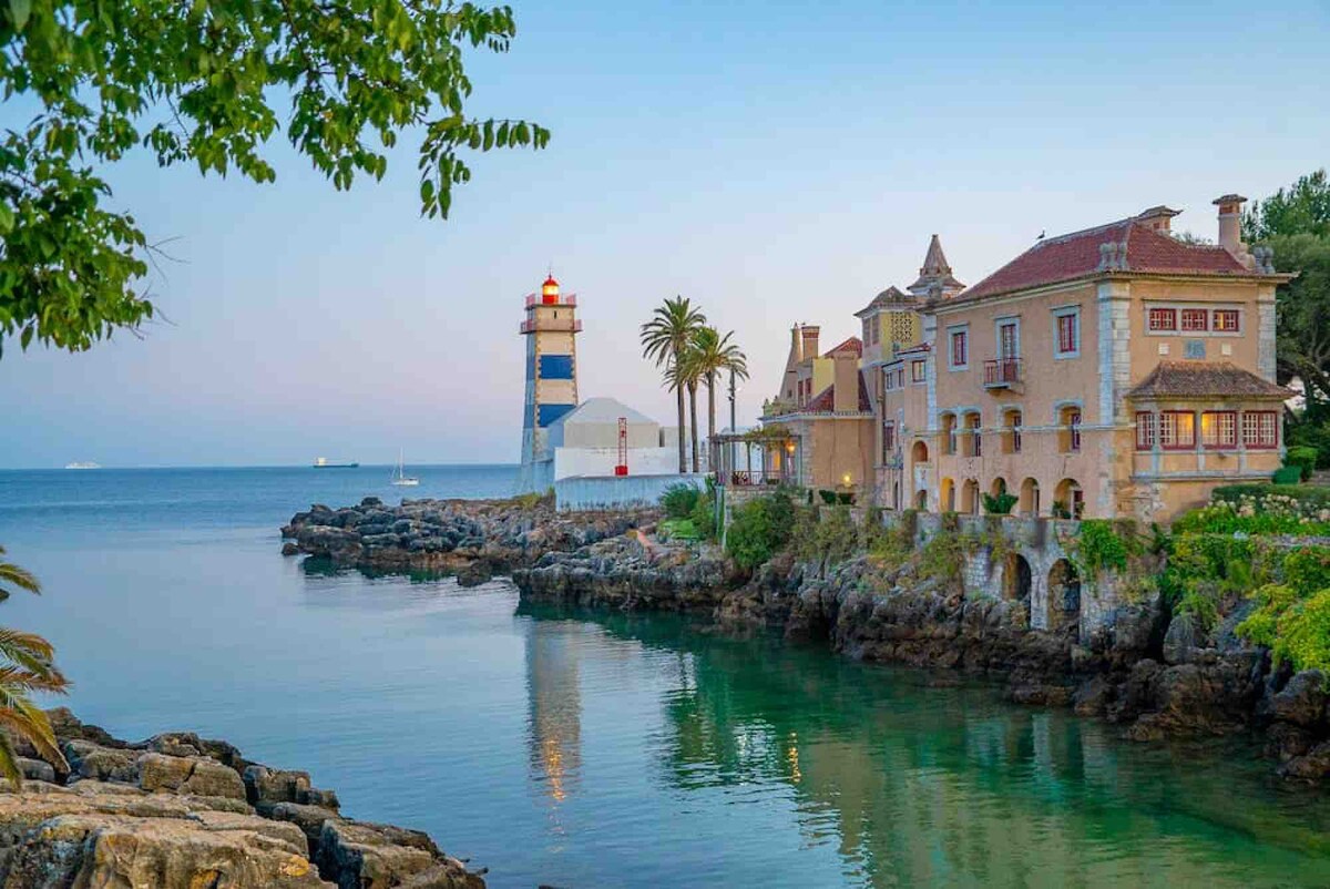 A scenic view shows a lighthouse beside a historic building with palm trees lining the coastline. Calm water reflects the structures and the light fading from the sky, creating a serene atmosphere. The rocky shore adds texture to the peaceful setting.