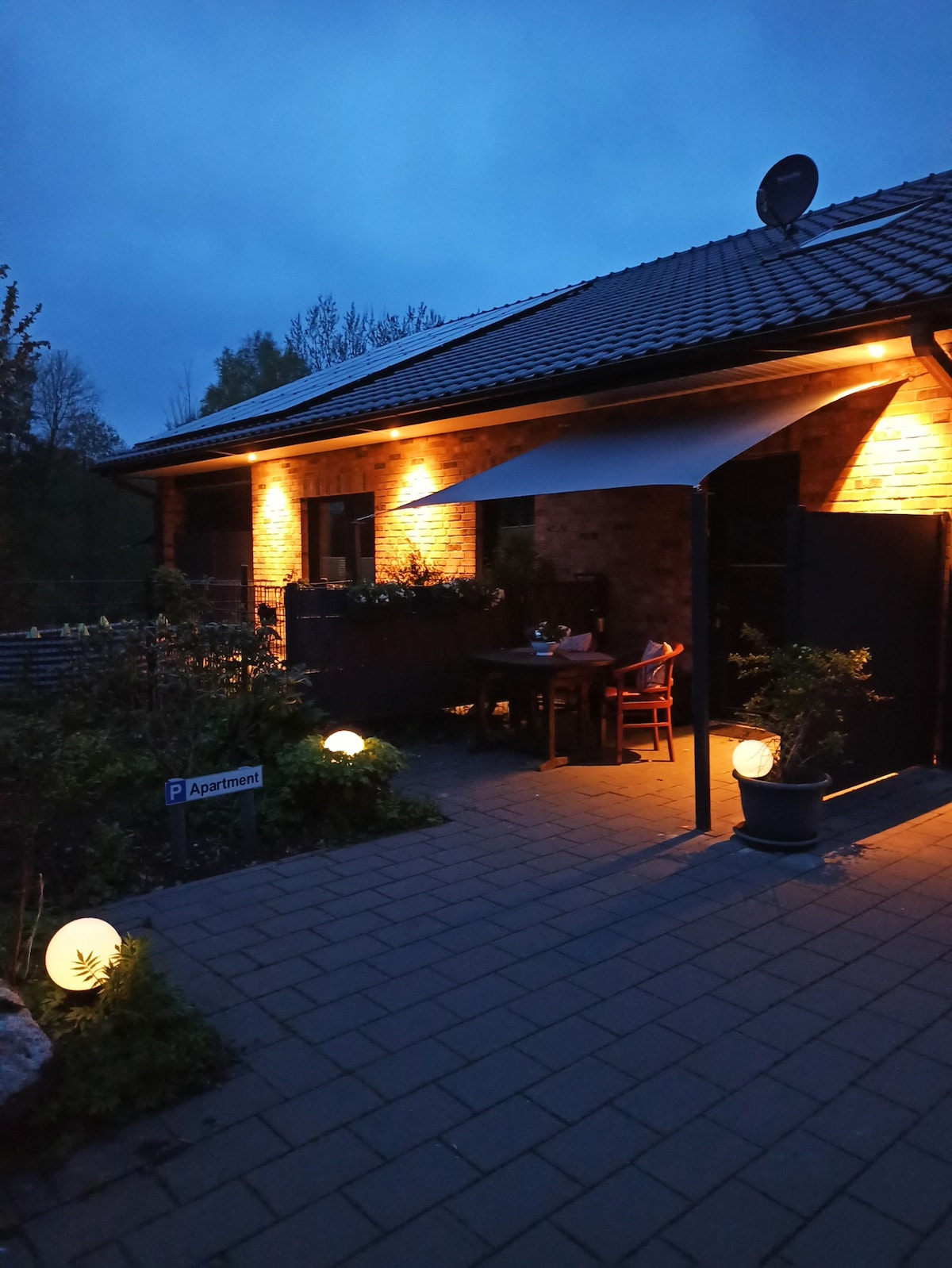 An outdoor terrace is softly illuminated by warm lights, creating a welcoming atmosphere. A wooden table with chairs is positioned under a canopy, surrounded by greenery. Decorative lanterns are placed on the ground, adding to the ambiance in the evening.
