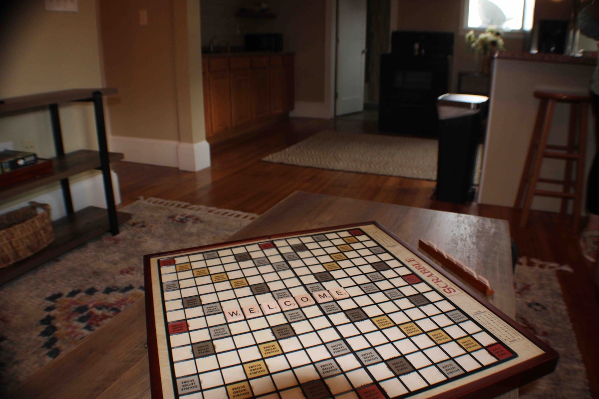 A classic board game is placed on a wooden coffee table in the foreground. The cozy living area features original hardwood floors and a welcoming layout, with the kitchen visible in the background, showcasing wooden cabinetry and essential appliances. Natural light softly illuminates the space.
