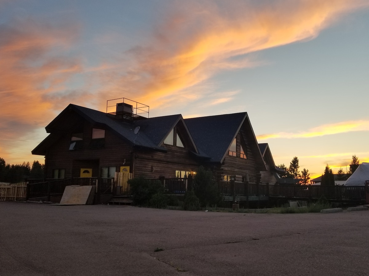 The wooden structure features a series of gabled roofs and large windows, set against a colorful sunset sky. The building is surrounded by open space, with decks visible along its exterior.