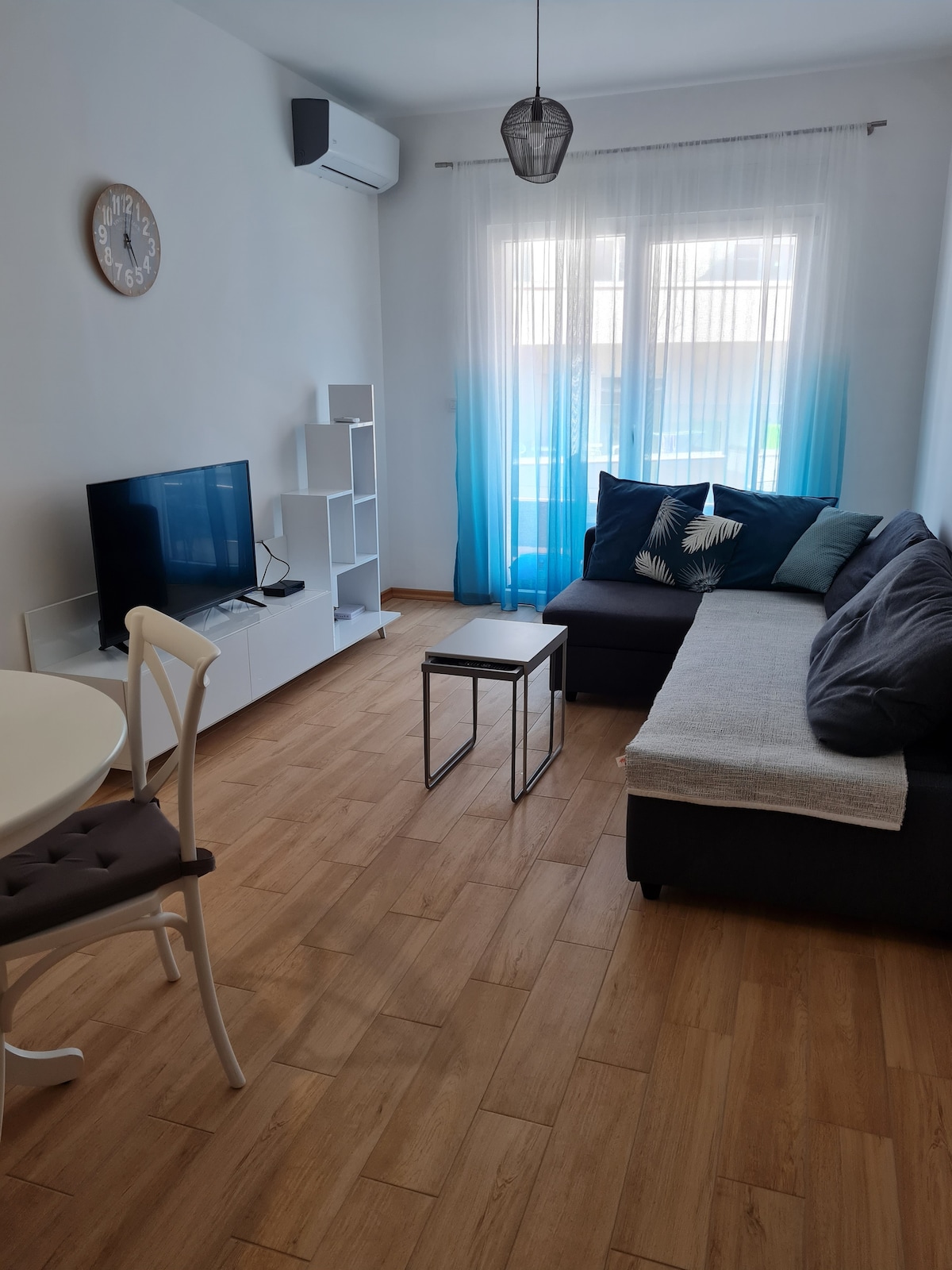 A cozy living area features a dark sectional sofa with decorative cushions, adjacent to a light wood dining table and chair. A television sits on a white shelving unit, while light curtains gently filter sunlight from a window.