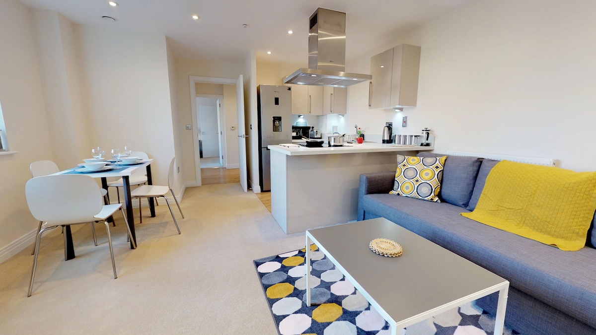 The open-concept living area features a modern kitchenette with stainless steel appliances. A grey sofa is paired with colorful cushions, and a coffee table sits on a patterned area rug. A dining table for four is visible, creating a welcoming space for guests.