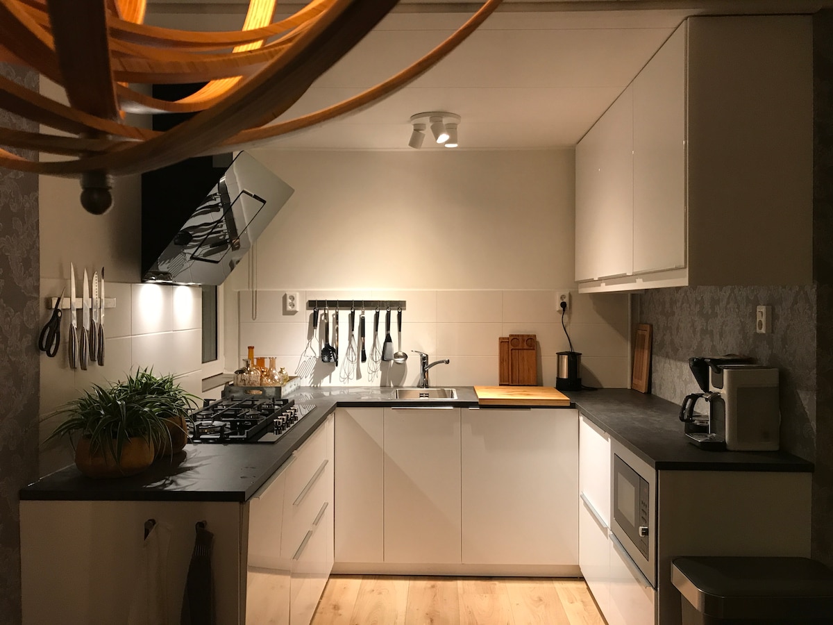 A modern kitchen features sleek white cabinetry and a dark countertop. Cooking utensils and knives are neatly arranged on the wall, while a gas stove and oven are integrated into the space. A small plant adds a touch of greenery, contributing to the functional and inviting ambiance.
