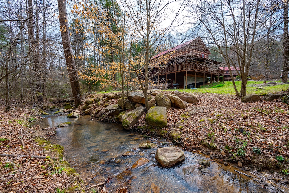A serene mountain creek flows gently past a log cabin, set against a backdrop of trees and a grassy area. The cabin features a rustic design with a covered porch. Autumn leaves are visible on nearby trees, adding a touch of color to the natural setting.
