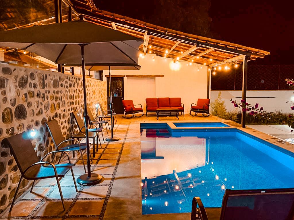 An inviting pool area with ambient lighting highlights a tranquil swimming pool surrounded by comfortable seating. Red couches and lounge chairs offer relaxation options, while a shaded umbrella provides respite from sunlight. The rustic stone wall adds character to the serene setting.