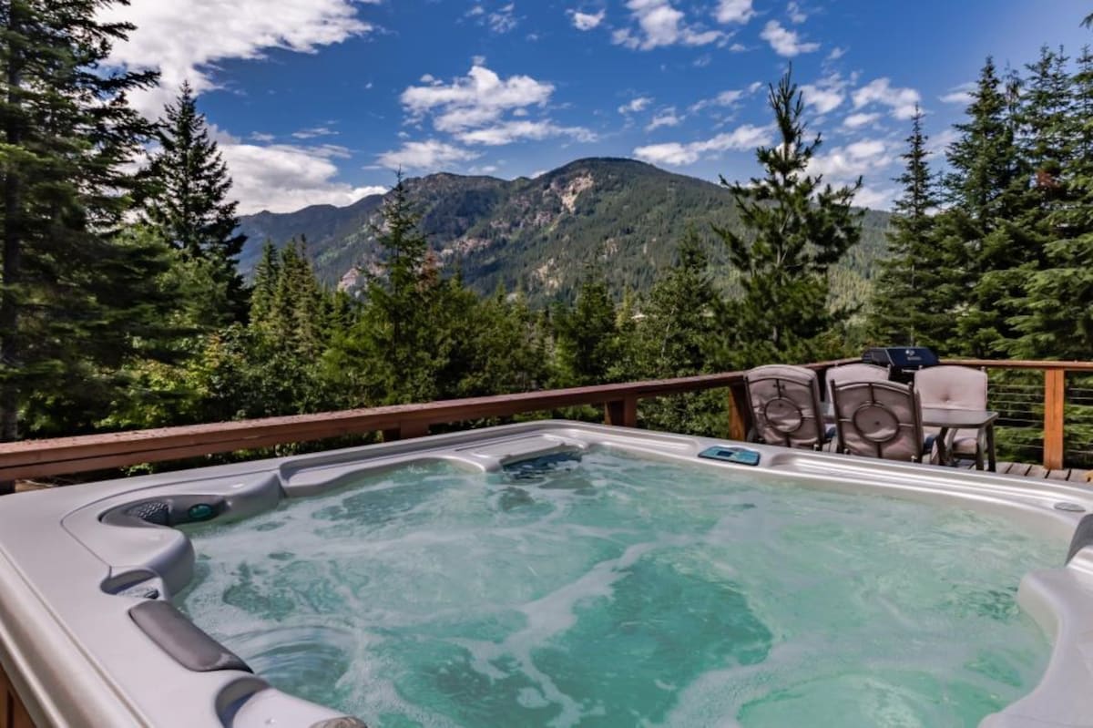 A private hot tub is situated on a wooden deck, surrounded by lush evergreen trees. Scenic mountain views are visible in the background under a partly cloudy sky, creating a tranquil outdoor setting. The hot tub features clear, bubbling water inviting relaxation.