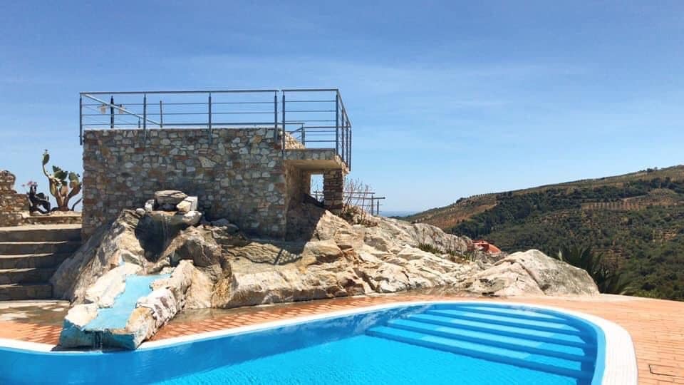 A stone structure with a railing is positioned beside a blue pool. The pool features a gradual entryway and is surrounded by a natural landscape of rolling hills. Clear skies and greenery are visible in the background, enhancing the tranquil outdoor setting.