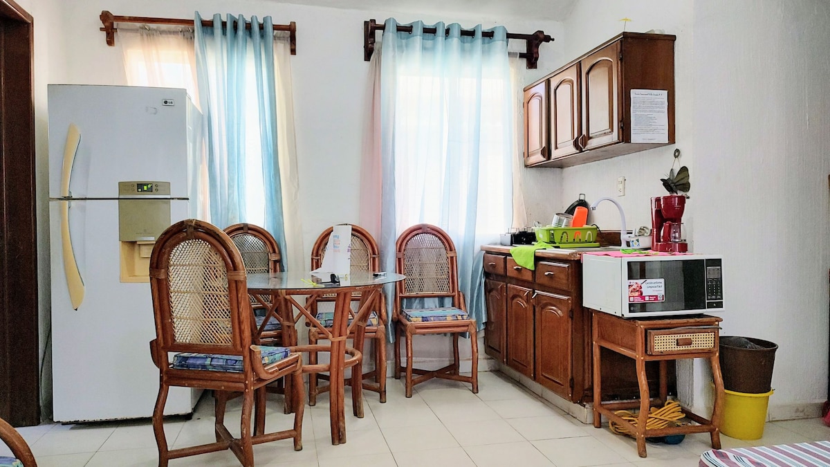 A well-lit kitchen area features wooden cabinets and a microwave, complemented by a refrigerator. A dining table with six chairs is present, with a colorful fruit bowl at its center. Natural light filters through sheer curtains, enhancing the inviting space.