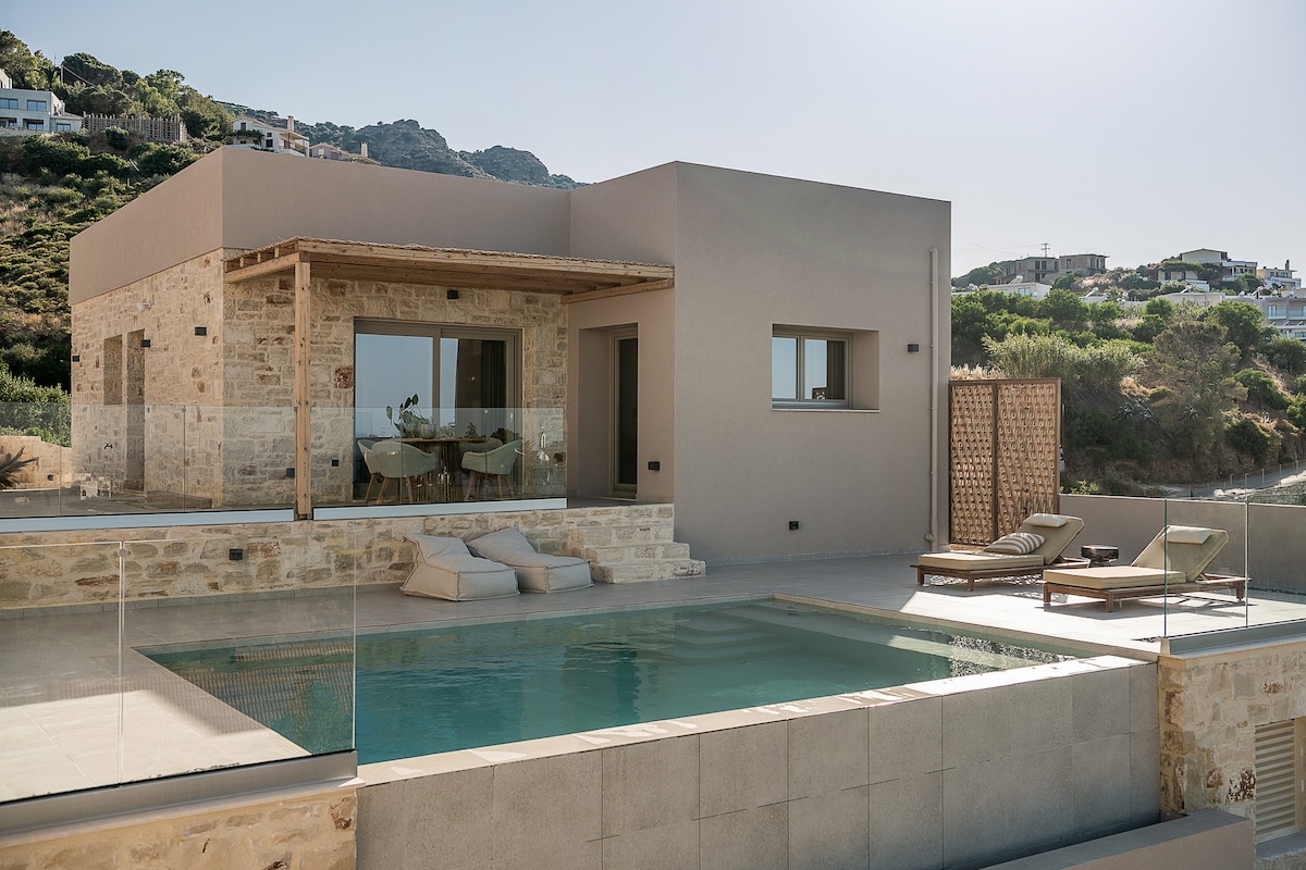 A modern villa is presented, featuring a private swimming pool with a stone deck and comfortable lounge chairs. The structure showcases a blend of natural stone and smooth walls, with large windows allowing for ample natural light. The surrounding hillside is visible in the background.