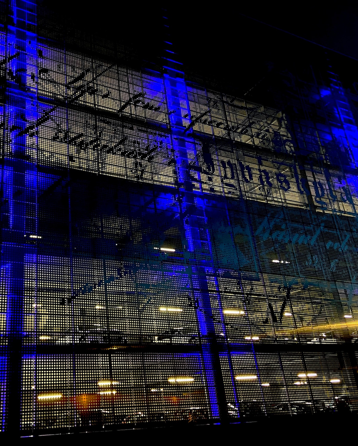 An illuminated contemporary building facade is visible at night, featuring intricate designs and vibrant blue lighting. The structure highlights detailed patterns and textures, complemented by warm light from the interior, creating a dynamic contrast against the dark sky.