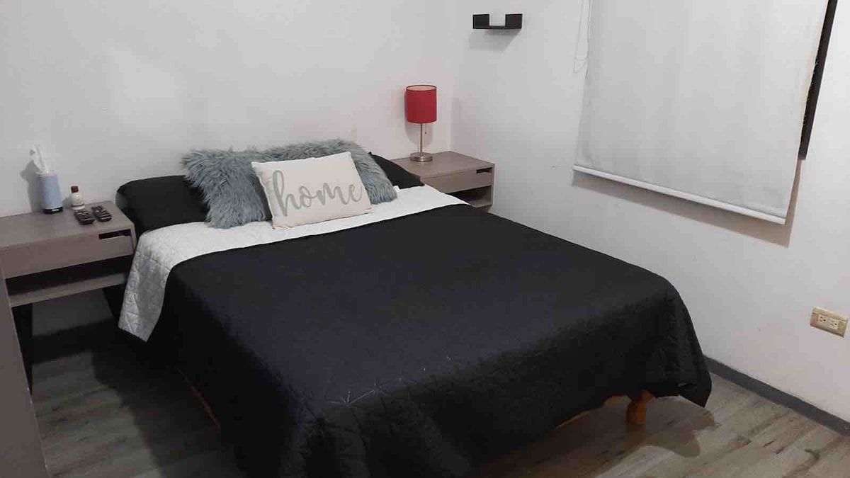A compact sleeping area features a spacious bed dressed in dark linens, complemented by a decorative pillow. Nightstands are positioned on either side, one holding a lamp and the other displaying various items. Natural light filters through a partially covered window, enhancing the serene ambiance.