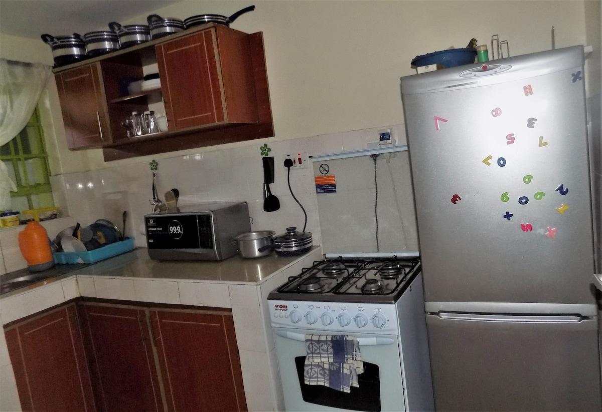 The kitchen is equipped with wooden cabinetry and a silver refrigerator adorned with colorful magnets. A gas stove is visible alongside a microwave and various utensils. Natural light enters through a nearby window, illuminating the countertops.