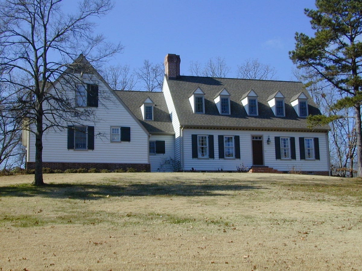 A charming reproduction style home is showcased, featuring a multi-gabled roof and symmetrical windows with shutters. The white exterior is contrasted by a natural landscape, with a large grassy area in the foreground and trees lining the property.