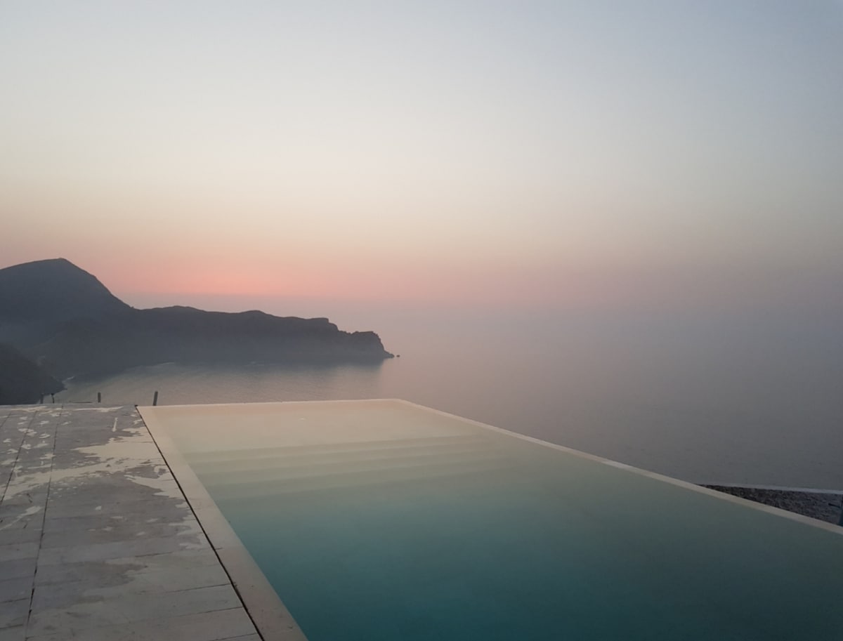 An infinity pool extends toward the horizon, creating a seamless connection with the tranquil sea. Soft twilight hues blend in the sky, while a distant mountainous coastline is visible, enhancing the serene atmosphere of the space.