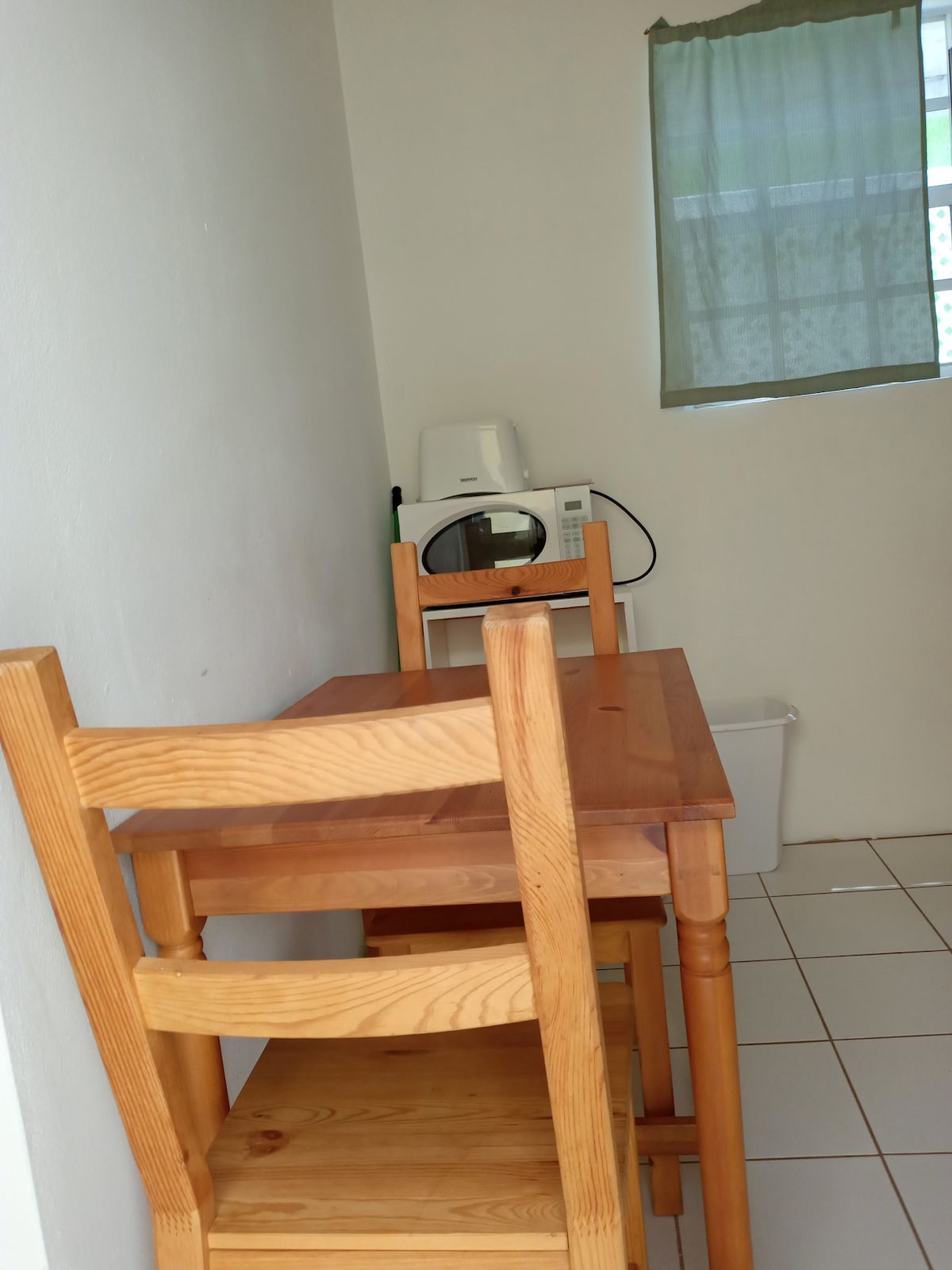 A simple kitchenette area features a wooden table with two matching chairs. A microwave and a small appliance are positioned on a countertop, while a waste bin is visible nearby. A light green curtain covers a window, allowing natural light to enter the space.