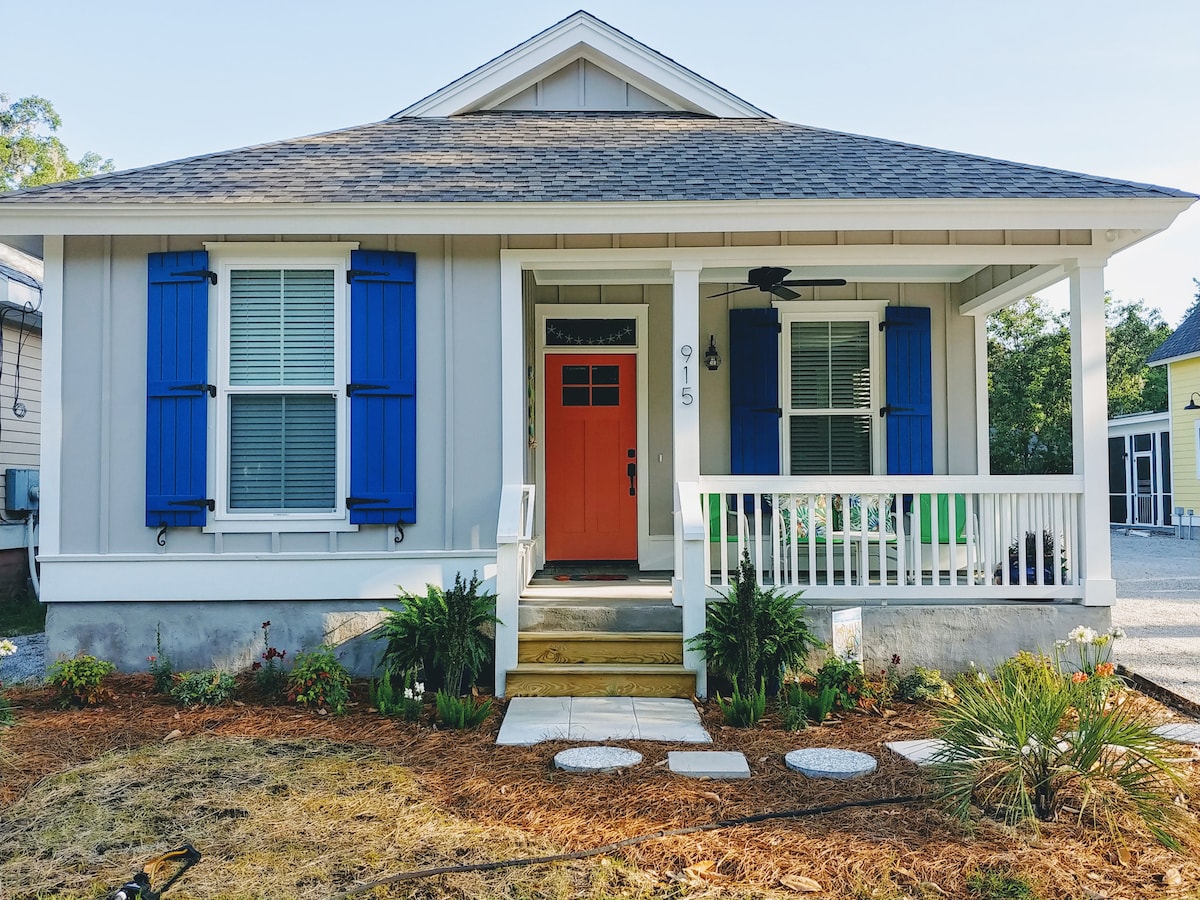 A newly built cottage features a welcoming front porch with a bright orange door framed by blue shutters. Lush landscaping surrounding the entrance includes palm and flowering plants, while a stone pathway guides visitors toward the steps. Ample natural light brightens the exterior.
