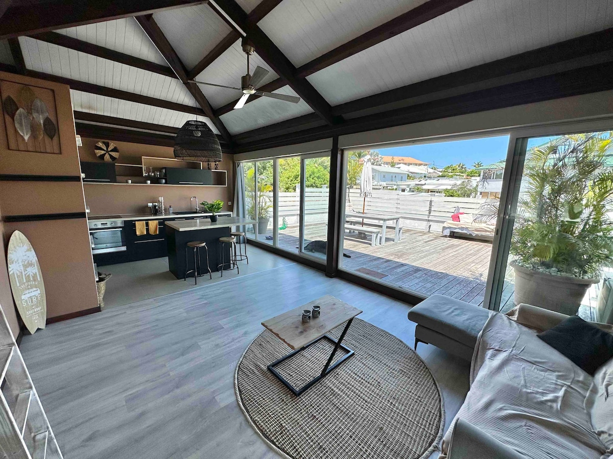 A spacious living area features large glass doors that open to a wooden terrace. A comfortable sofa and a circular rug are present, contributing to the relaxed ambiance. The kitchen area is visible, showcasing modern appliances and seating at a dark table. Light fills the space.