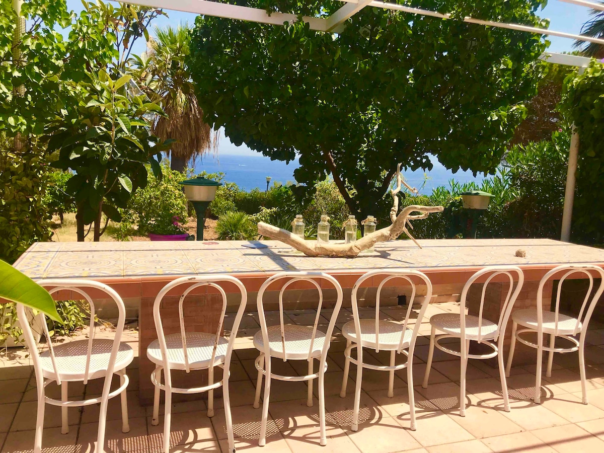 An outdoor dining area features a long, tiled table surrounded by six white chairs. Lush greenery and palm trees enhance the natural setting, while a glimpse of the sea can be seen in the background, providing a serene and inviting atmosphere.