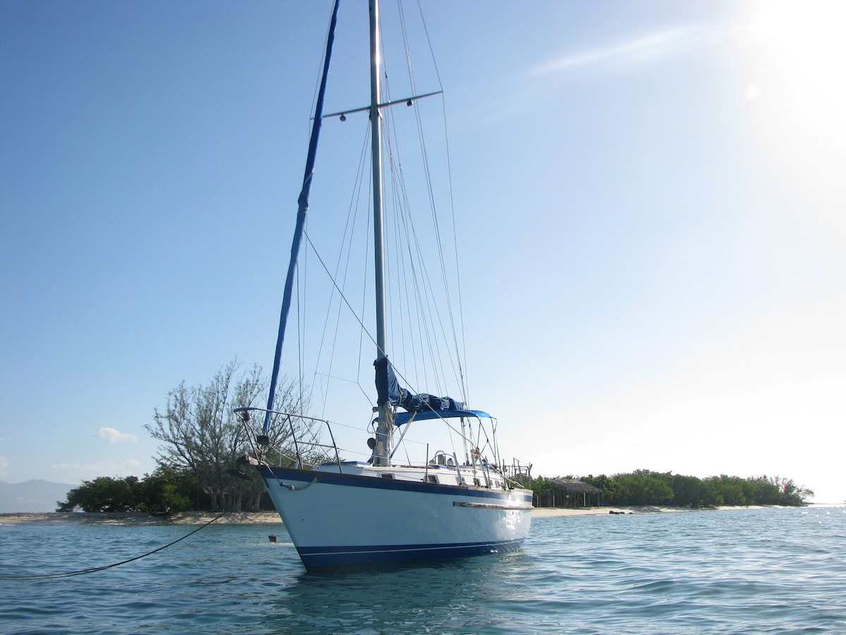 A well-maintained yacht is anchored in clear turquoise water, showcasing a tall mast and blue sails. The serene scene is complemented by a distant view of lush greenery and mountainous terrain under a bright sky.