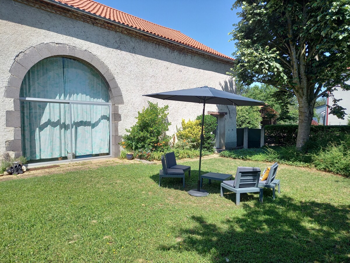 A private garden area is shown with a shaded seating arrangement that includes four chairs and a small table. Lush greenery surrounds the patio, and a large arched window is visible on the nearby building. The atmosphere is calm and inviting.