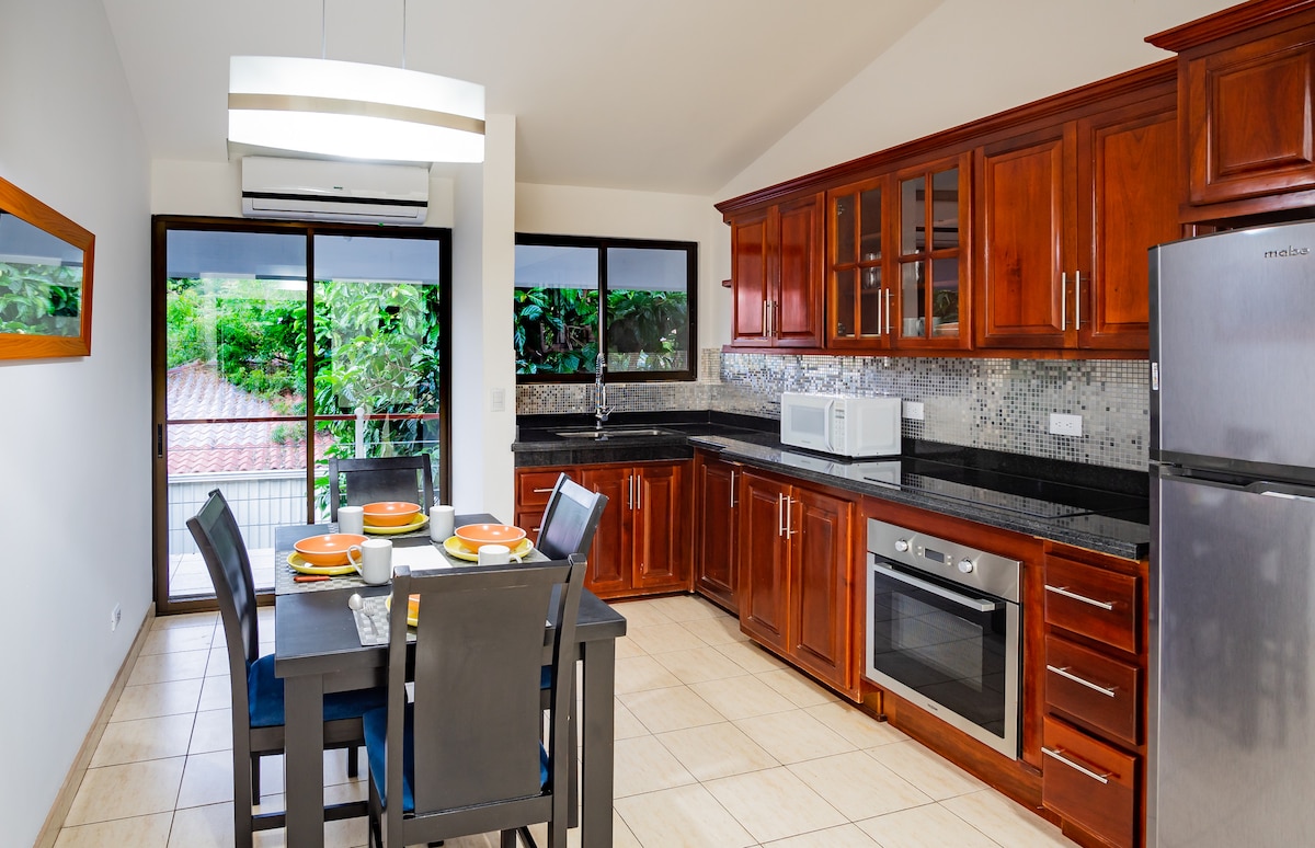 A modern kitchen is highlighted with rich wooden cabinetry and black countertops. The space features stainless steel appliances, including a microwave and oven. A dining area with a table set for four is visible, along with large windows offering views of lush greenery.
