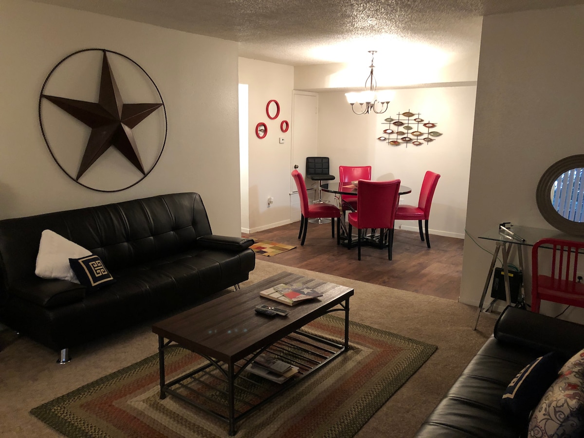 The living area features a black leather sofa and a second seating arrangement, complemented by a coffee table. A dining space with red chairs surrounds a circular table. Wall decor includes a large star and metallic wall art, creating a stylish atmosphere.