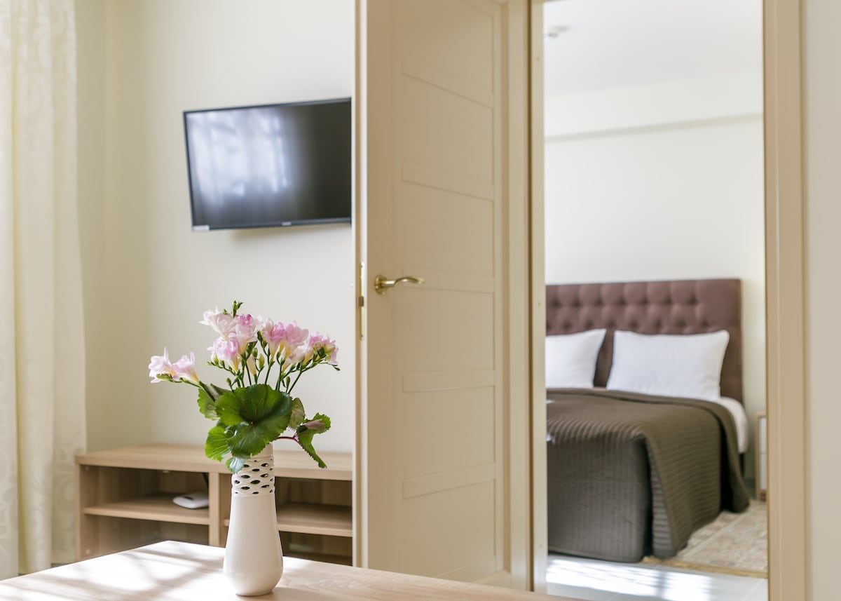 A view through an open door reveals a separate bedroom featuring a large double bed with neutral bedding. A flat screen TV is mounted on the wall in the living area. A vase with pink flowers is placed on a light-colored tabletop, adding a touch of color.