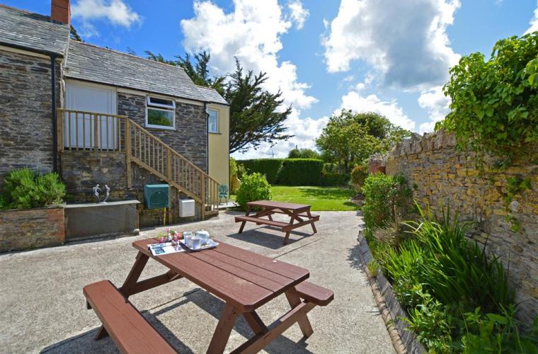 An enclosed private garden area features two picnic tables surrounded by well-maintained grass and planted greenery. The stone walls add a rustic touch, and the bright blue sky with fluffy clouds creates a cheerful atmosphere. Nearby, a cottage staircase leads to an upper level.
