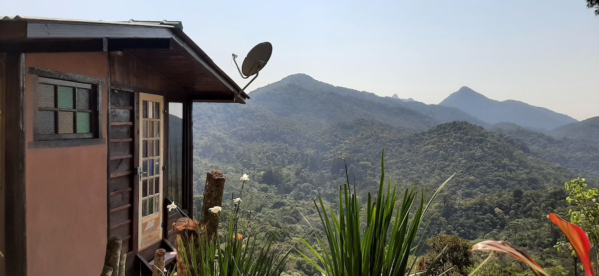 The exterior of a rustic mountain chalet is shown, with wooden accents and a satellite dish. Lush greenery and rolling hills extend into the distance, underscoring the serene natural setting. The area is bathed in natural light, creating a tranquil atmosphere.