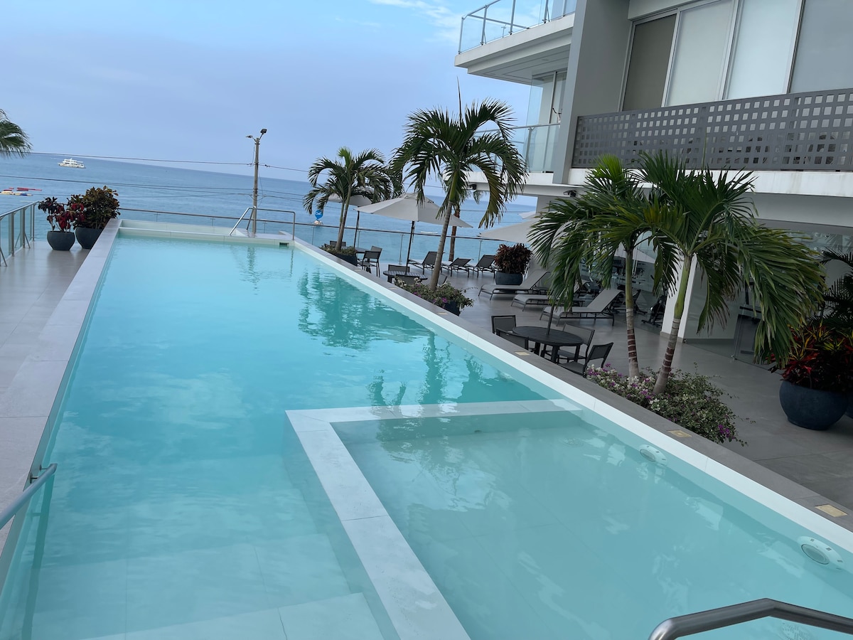 A refreshing outdoor pool stretches alongside the apartment, with clear turquoise water inviting relaxation. Palm trees are positioned around the area, and lounge chairs are set up on a sunlit deck. The ocean is visible in the background, enhancing the serene coastal ambiance.