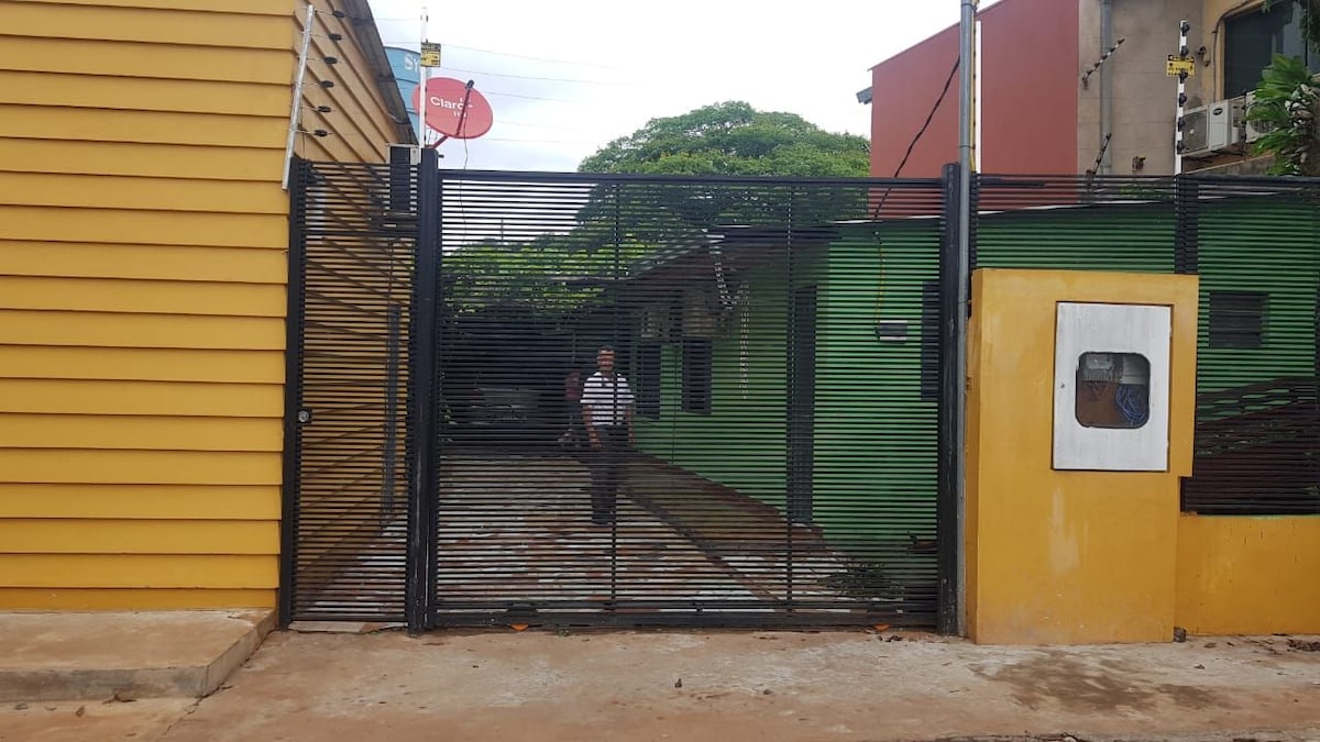 A metal gate stands open, revealing a spacious courtyard lined with cobblestones. Two buildings are visible, one painted in green and the other in yellow, surrounded by greenery. A satellite dish is mounted on the yellow building, enhancing the visible outdoor area.